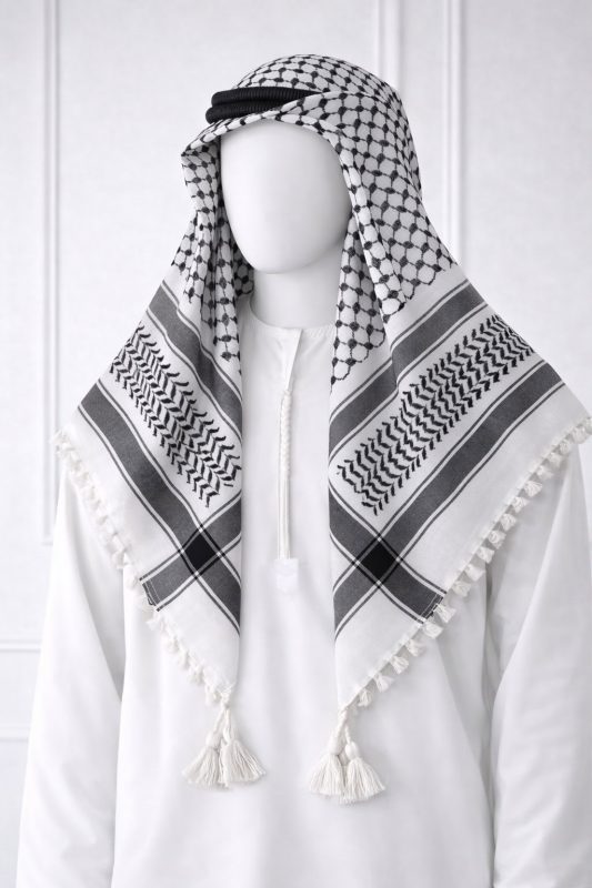 Classic white keffiyeh shemagh scarf featuring black geometric patterns and tassel edges worn over a white thobe for a traditional Arabic look.
